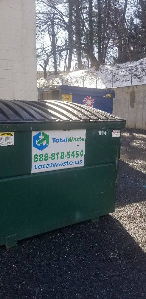 A green commercial dumpster from Total Waste in a parking area with another blue dumpster in Baltimore, MD.