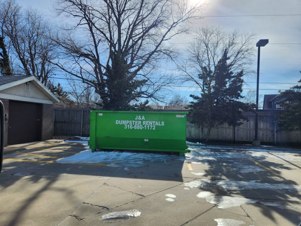 A green J & A Haul Away & Dumpster Rentals dumpster in a parking lot with patches of snow in Derby, KS.