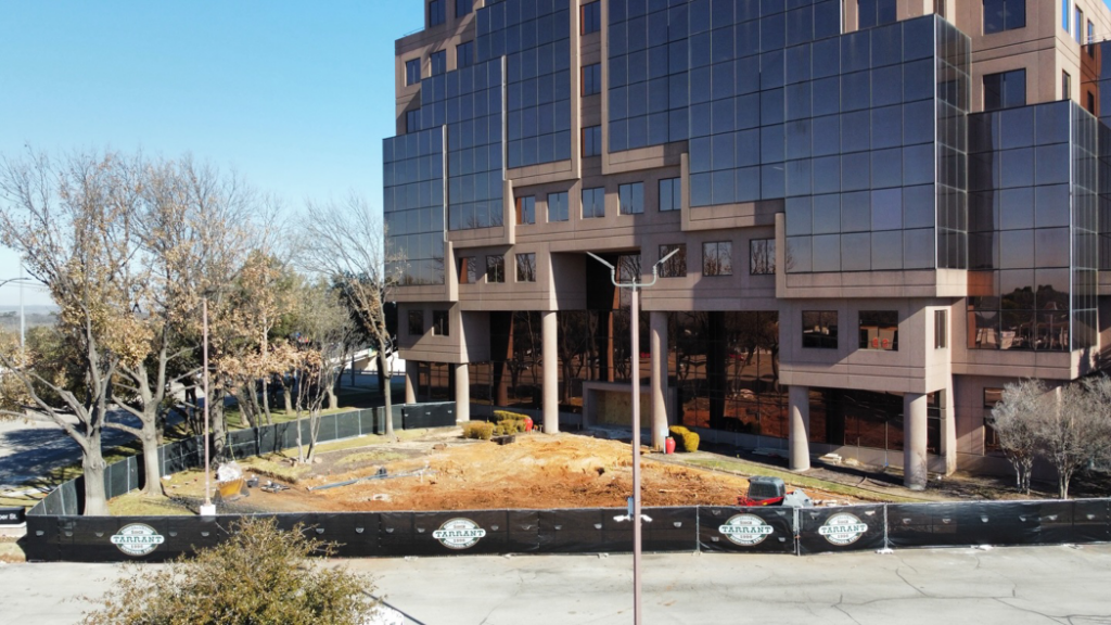 A ground-level view of a commercial construction site with a branded fence and equipment by Tarrant Construction Services, LTD in Fort Worth, TX