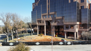 A ground-level view of a commercial construction site with a branded fence and equipment by Tarrant Construction Services, LTD in Fort Worth, TX