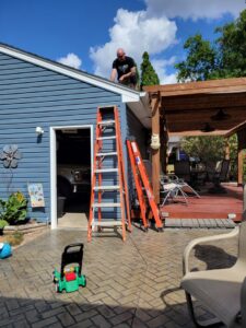 A handyman on a roof next to a gutter, with ladders leaning against the house, performing cleaning or repair by Cedar Valley Maintenance and Handyman Services LLC in Waterloo, IA.
