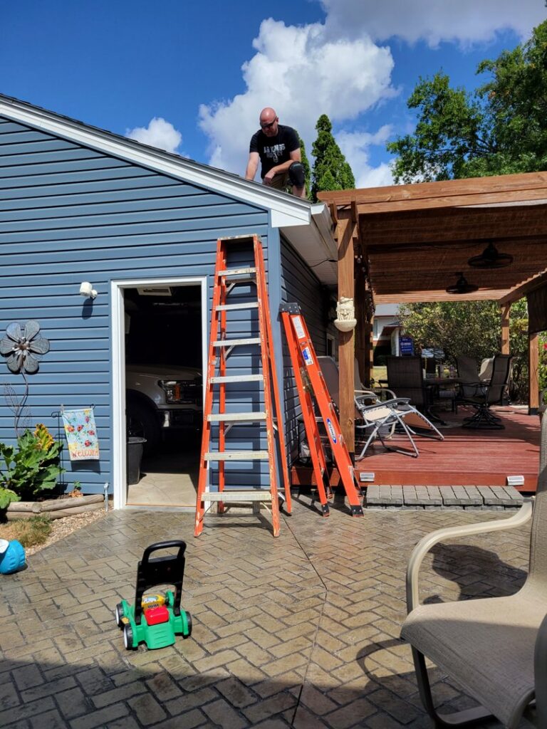 A handyman on a roof next to a gutter, with ladders leaning against the house, performing cleaning or repair by Cedar Valley Maintenance and Handyman Services LLC in Waterloo, IA.