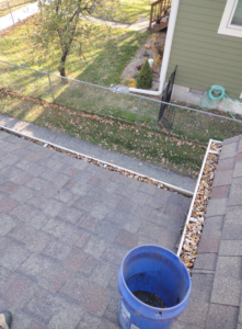 A blue bucket on a roof next to a gutter full of leaves, illustrating gutter cleaning services by Schu Can Do 2-Property Services LLC in Warren, MI.