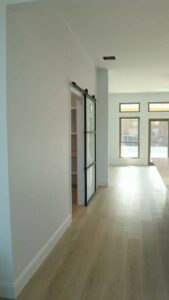 A renovated hallway with light wood flooring and a stylish black-framed barn door leading to a pantry, by Whole House Remodel & Design North Dallas Tx in Frisco, TX.
