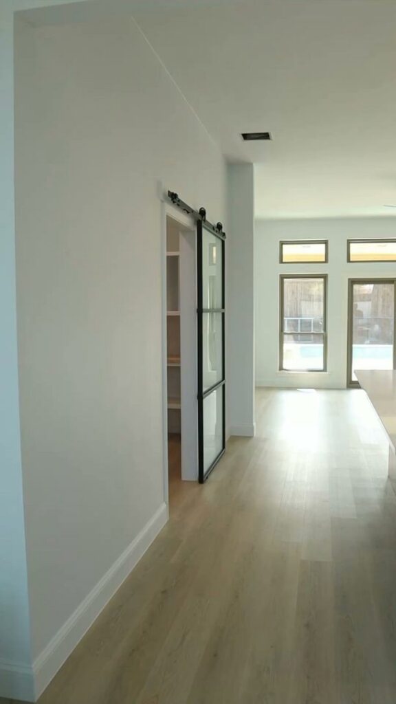 A renovated hallway with light wood flooring and a stylish black-framed barn door leading to a pantry, by Whole House Remodel & Design North Dallas Tx in Frisco, TX.