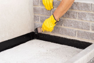 A handyman applying black waterproofing sealant to a concrete corner for Tito's Home Improvement LLC in Stamford, CT.