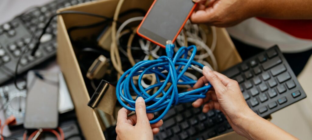 Hands sorting tangled cables and a smartphone into a cardboard box for e-waste recovery by E-Waste Recovery Systems in Sacramento, CA.