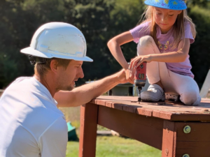 A handyman from Woods Builders in Grand Rapids, MI, guiding a child using a drill to assemble a wooden playset.