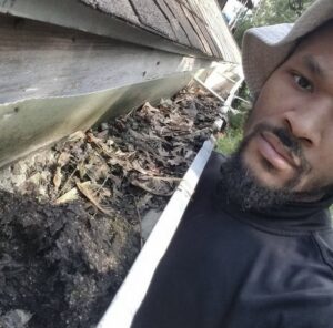 A handyman from Applied Pressure LLC in Detroit, MI, cleaning a gutter filled with leaves and debris.