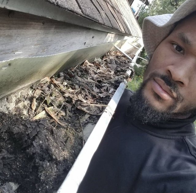A handyman from Applied Pressure LLC in Detroit, MI, cleaning a gutter filled with leaves and debris.