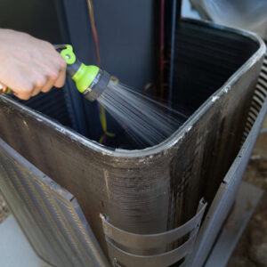 A handyman cleaning an outdoor AC unit coil with a hose, a maintenance service by AC Services of Minden in Minden, LA.