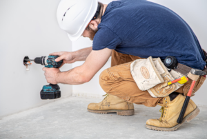 A handyman wearing a hard hat and tool belt drilling an electrical outlet, offering services from Handyman Services of Albuquerque in Albuquerque, NM.