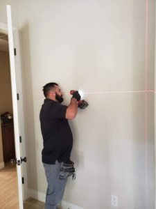 A handyman using a drill on a wall with a laser level for precise installation, performed by NH Handyman in San Diego, CA.