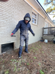 A handyman in a work jumpsuit inspecting a crawl space opening at a residential property by BearHome Service in Mount Pleasant, SC