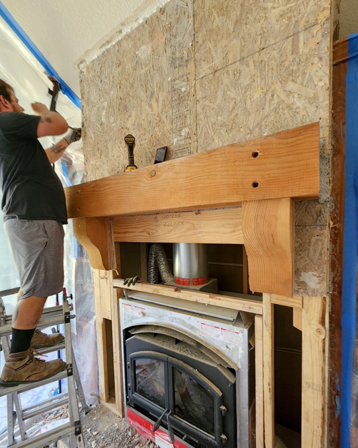A handyman on a ladder installing a wooden mantel and framing around a fireplace insert for Portland Fireplace and Chimney Inc. in Portland, OR.