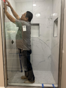 A handyman installing a glass shower door in a tiled bathroom for Saul Perez Renovations LLC in Macon, GA.