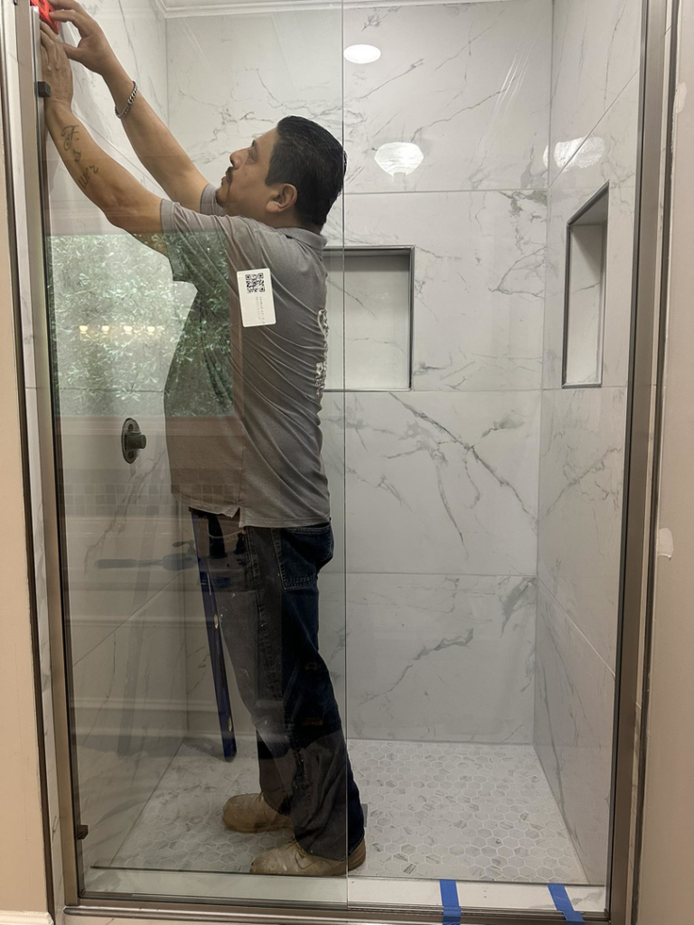 A handyman installing a glass shower door in a tiled bathroom for Saul Perez Renovations LLC in Macon, GA.