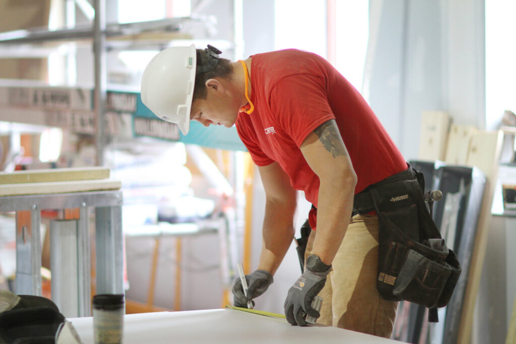 A handyman wearing a hard hat and tool belt measuring material with a tape measure for Cheever Builders in Grand Rapids, MI.