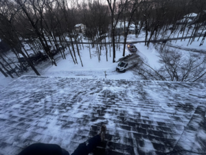 A handyman on a snowy roof with a work van parked below, performing winter roof services for BETHO Construction INC in Milford, MA.