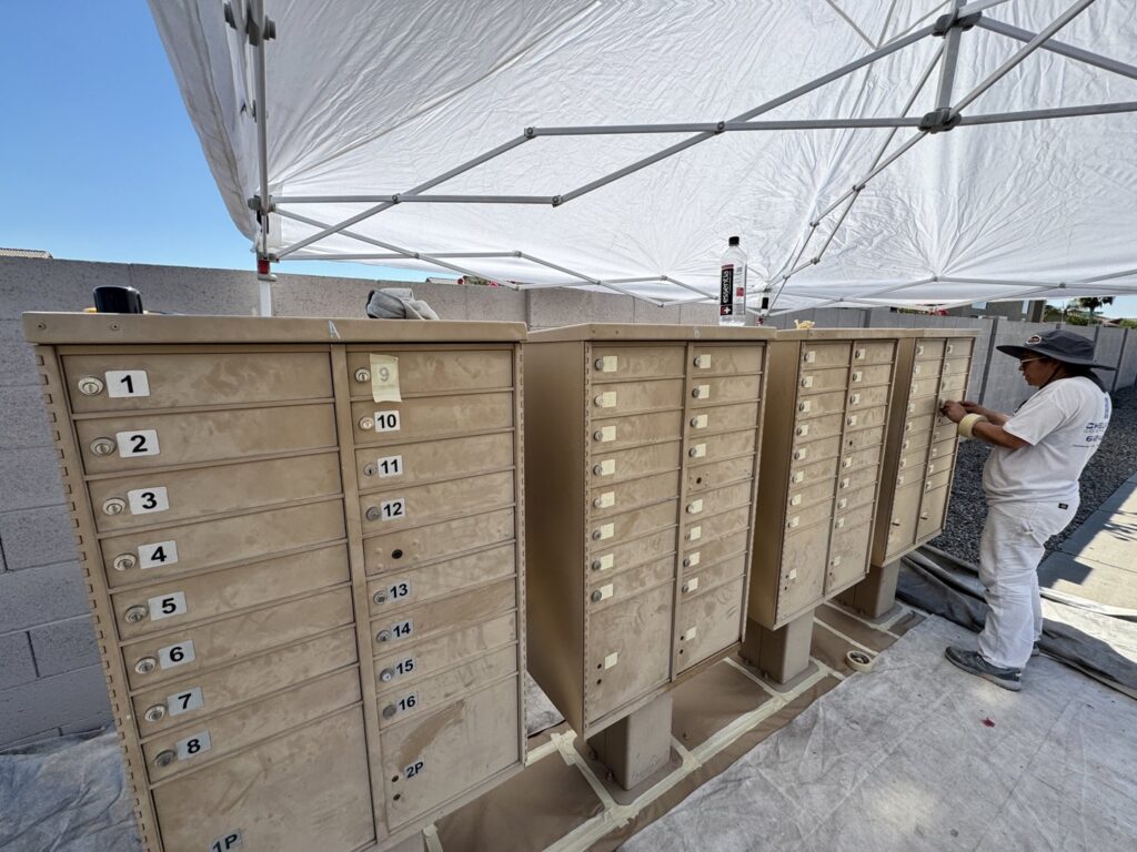 A handyman from State 48 Painting Company LLC painting community mailboxes under a canopy in Queen Creek, AZ.