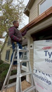 A handyman repairing a gutter or fascia board while on a ladder for Cedar Siding Repair in Centerville, OH.