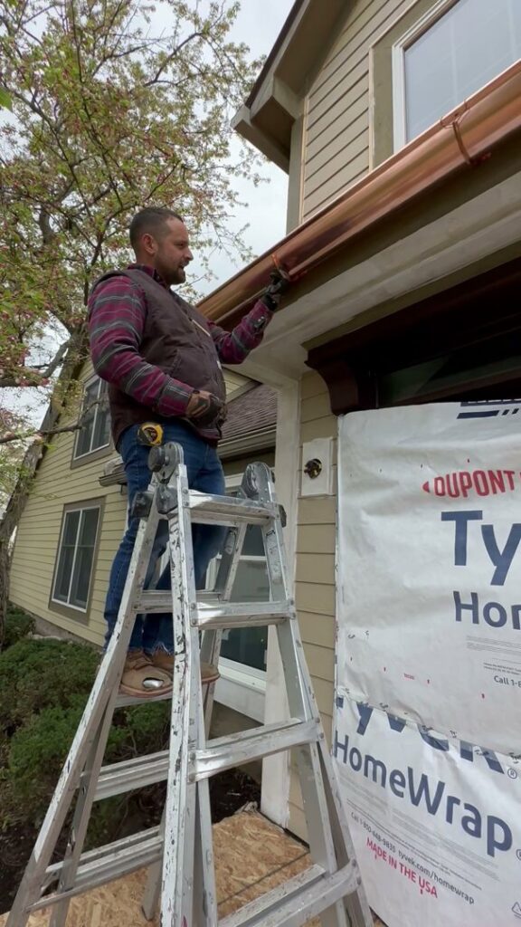 A handyman repairing a gutter or fascia board while on a ladder for Cedar Siding Repair in Centerville, OH.