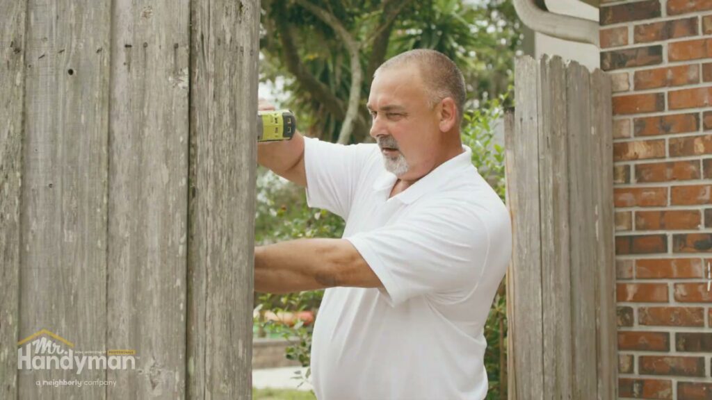 A handyman repairing a wooden fence with a drill, a service provided by Mr. Handyman of Olathe, Gardner in Olathe, KS.