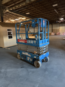 A Handyman branded scissor lift inside a building with a shed in the background, available at Handyman Rental in Portland, ME.