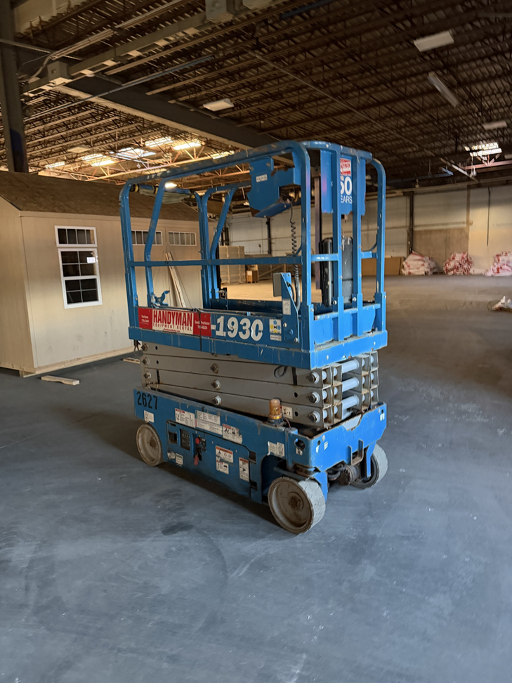 A Handyman branded scissor lift inside a building with a shed in the background, available at Handyman Rental in Portland, ME.