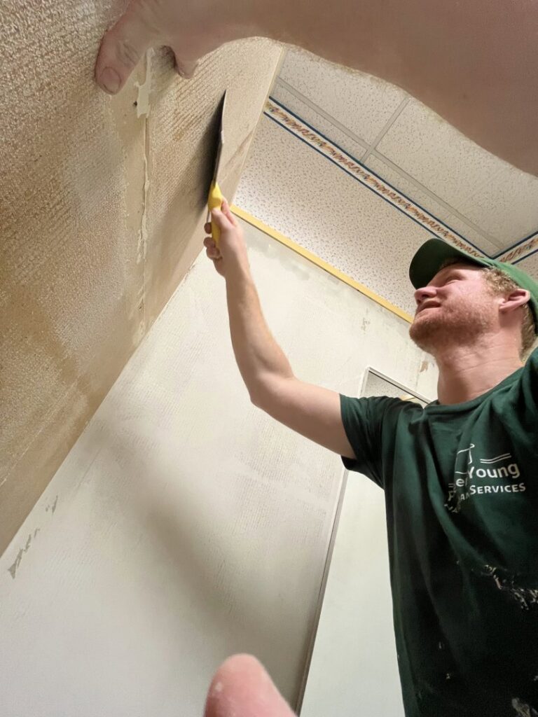 A handyman from Forever Young Handyman Services in Boise, ID, actively scraping wallpaper off a wall.