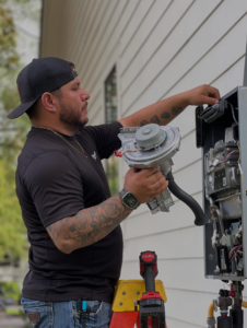 A handyman servicing an outdoor tankless water heater with a component in hand for Sandobros Plumbing in Charleston, SC