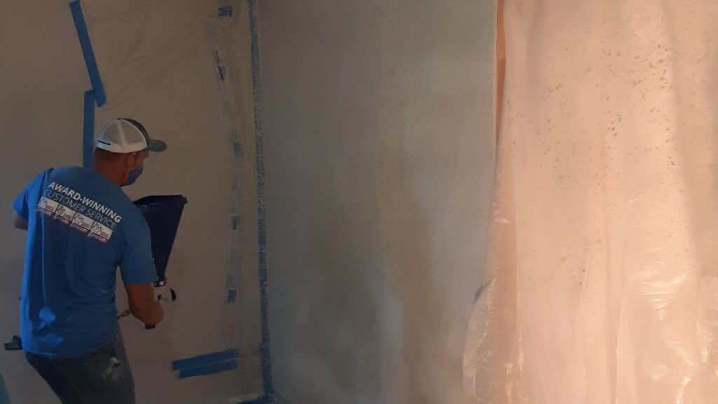 A handyman spray painting a wall, with protective plastic sheeting, showcasing painting services by Homefix Lawrence in Lawrence, KS.