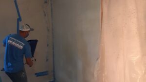 A handyman spray painting a wall, with protective plastic sheeting, showcasing painting services by Homefix Lawrence in Lawrence, KS.