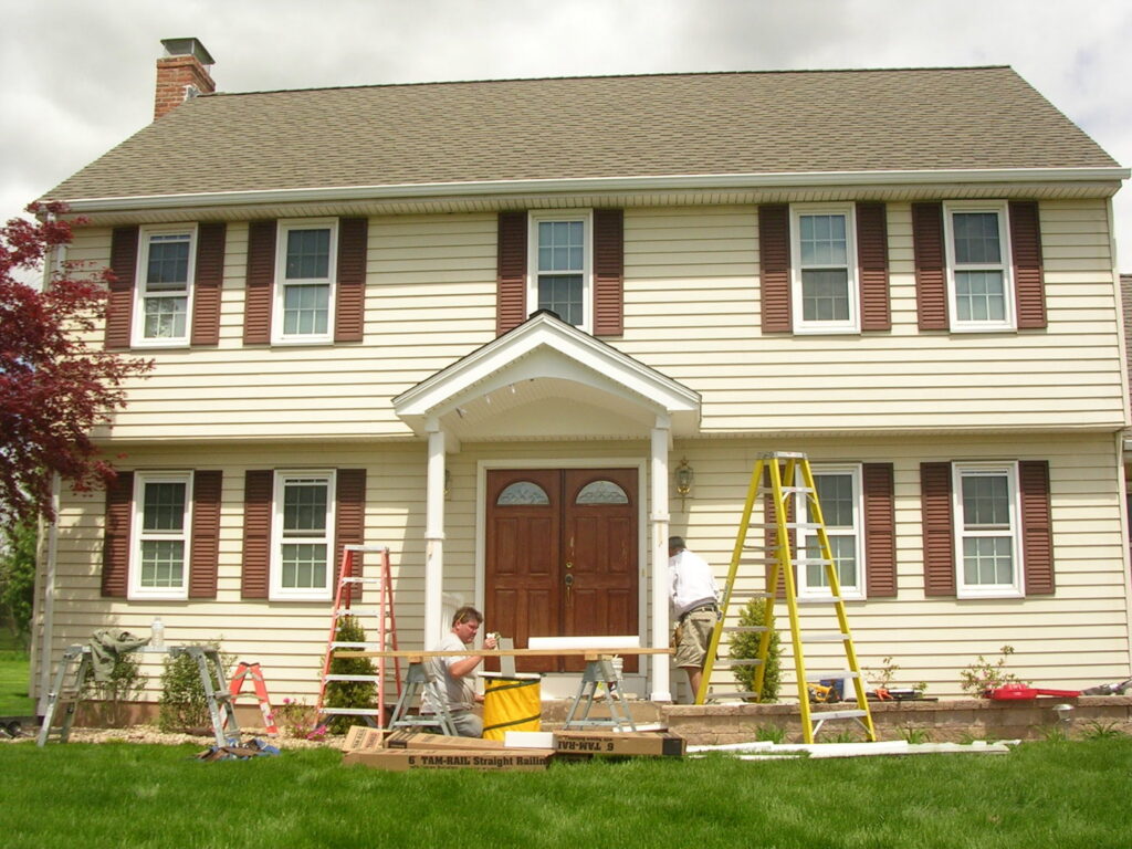Two handymen installing trim on the front of a house, with ladders and tools, by Bartlett Brainard Products Co., Inc. in West Hartford, CT.