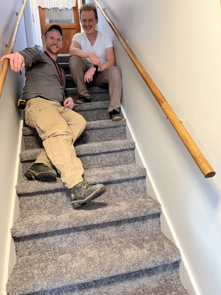 Two handymen sitting on a newly carpeted staircase with a wooden handrail, showcasing completed work by Forever Young Handyman Services in Boise, ID.