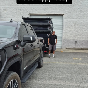 A happy man stands beside a black pickup truck towing stacked roll-off containers, ready for junk removal with Lewis Outdoor Services LLC in Augusta, GA.