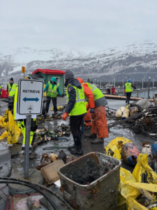 A harbor cleanup crew collecting debris and junk into yellow bags with snowy mountains, supported by Alaska Waste in Anchorage, AK.