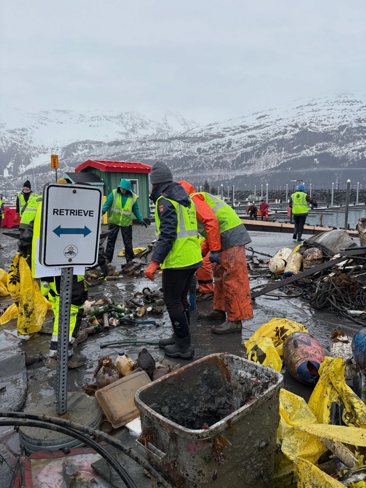 A harbor cleanup crew collecting debris and junk into yellow bags with snowy mountains, supported by Alaska Waste in Anchorage, AK.