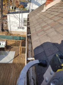 A heavily clogged gutter filled with leaves and debris, seen from a ladder next to a wooden deck, ready for cleaning by Schu Can Do 2-Property Services LLC in Warren, MI.