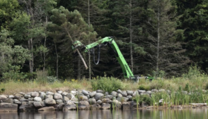 A green heavy equipment machine with a grapple attachment removing a tree trunk near water for DJ's Tree Service in Colchester, VT.
