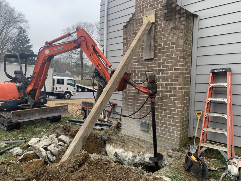 An excavator and helical pier installation in progress next to a brick chimney by Ram Jack Foundation Solutions in Hampton, VA.