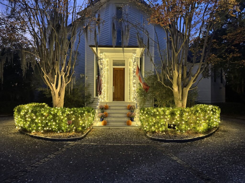 A charming historic house with white lights illuminating the porch and surrounding bushes by Lights Made Easy of Charleston, SC