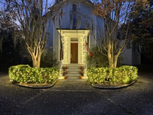 A charming historic house with white lights illuminating the porch and surrounding bushes by Lights Made Easy of Charleston, SC