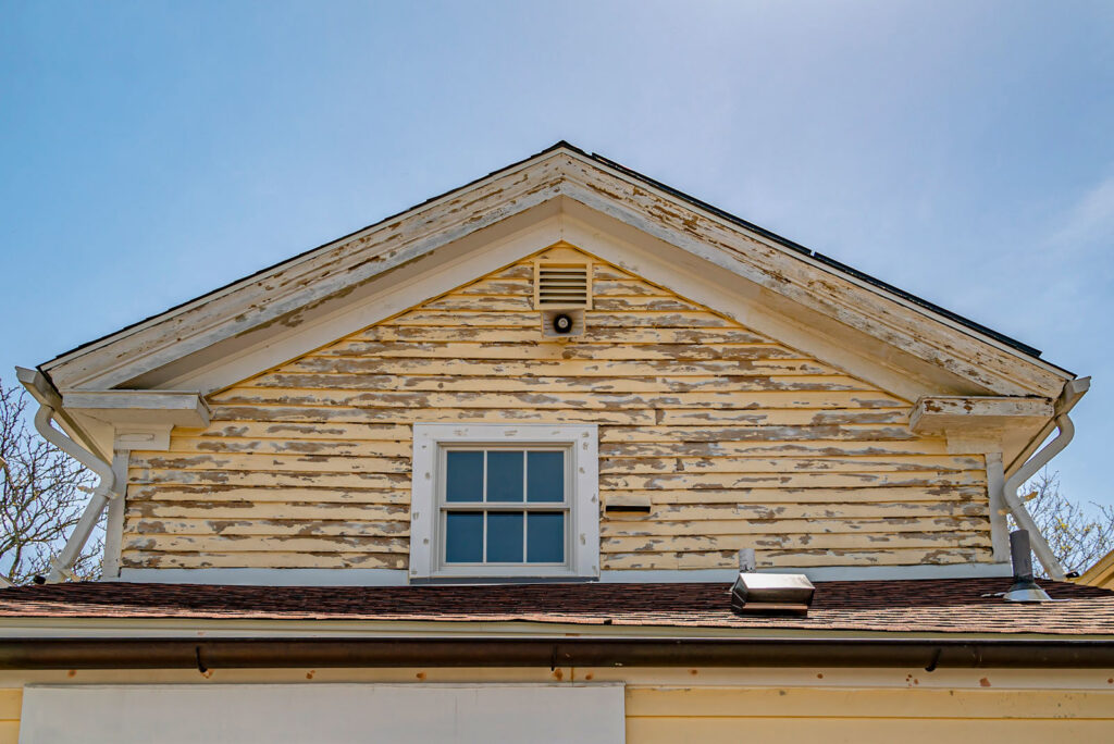 The gable end of a house with severely peeling yellow paint, showing a need for GO Painting's handyman services in New Haven, CT