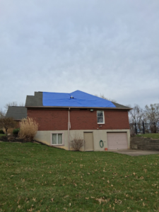 A house with a blue tarp covering a damaged roof, indicating repair work by Elegant Home Exteriors in Flourance, KY.