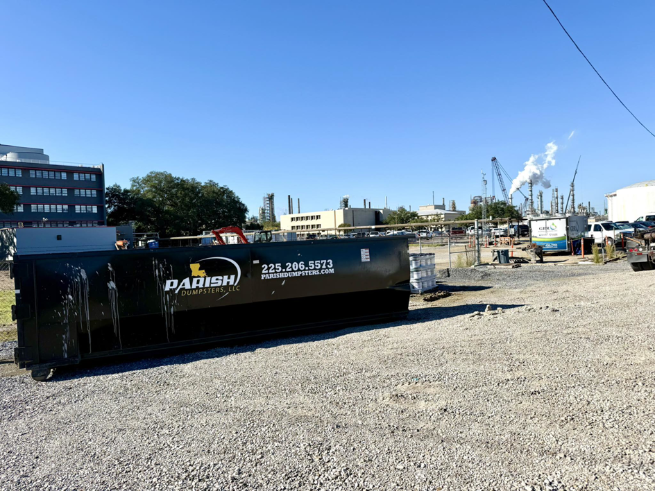 An industrial dumpster from Parish Dumpsters, LLC placed at a gravel lot near a factory in Baton Rouge, LA.