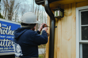 A worker installing a dark downspout on a wooden-sided house for The Brothers that just do Gutters in Sioux Falls, SD.
