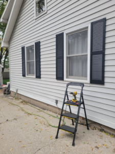 A handyman installing new black shutters on a house exterior, with a drill on a ladder, by Cedar Valley Maintenance and Handyman Services LLC in Waterloo, IA.