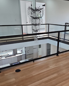 A second-floor interior balcony with stylish glass railings overlooking a modern kitchen, installed by Alpha Railing Company in Tacoma, WA.