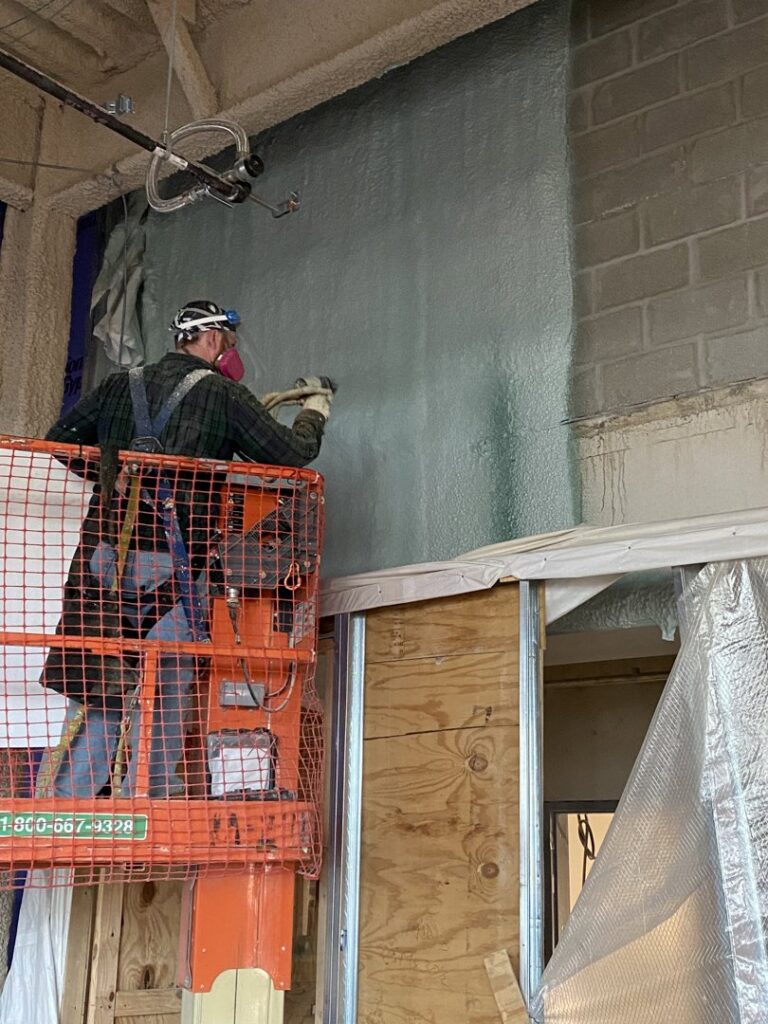 A worker on a boom lift applying spray foam insulation to an interior commercial wall, part of a general contractor's project by Olympus Spray Foam in Philadelphia, PA.
